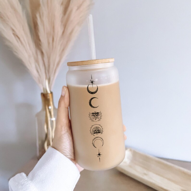 May include: A frosted glass reusable cup with a bamboo lid and a straw. The cup has a black and white moon phase design with stars.