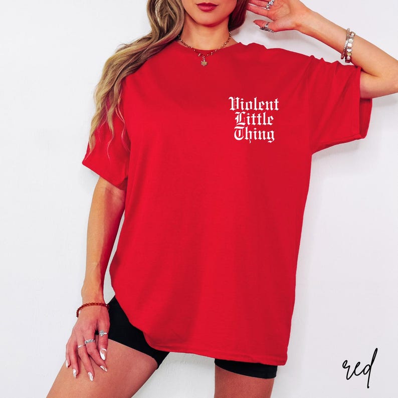 May include: A vibrant red t-shirt with the text "Violent Little Thing" in white gothic lettering on the upper left chest. The shirt is a solid color and has short sleeves.