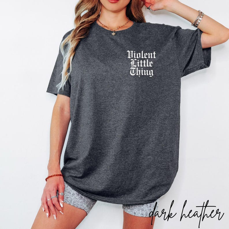 May include: Dark heather gray t-shirt with the text "Violent Little Thing" in white gothic font on the upper chest. The shirt is a short-sleeved, casual style.