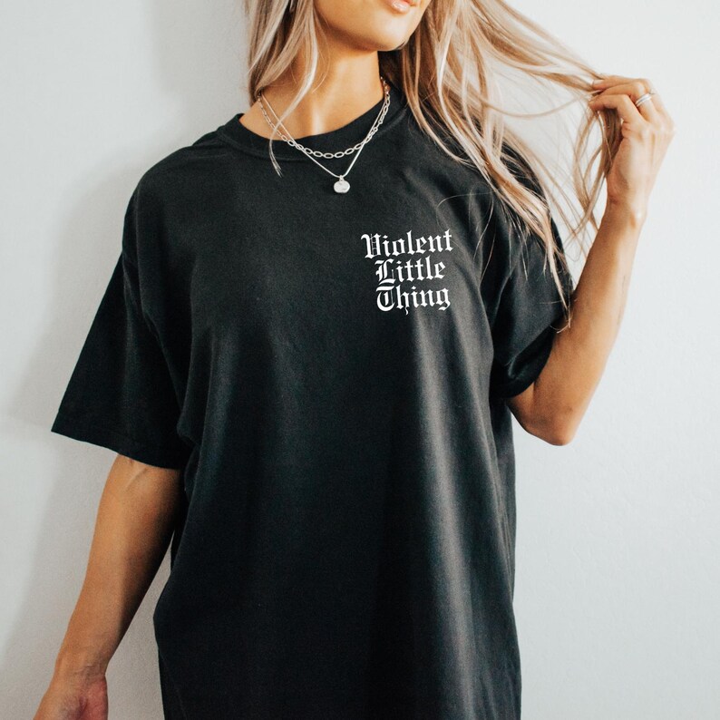 May include: Black t-shirt with the text "Violent Little Thing" in white gothic font on the upper left chest. The shirt has short sleeves and a relaxed fit. A silver necklace is visible.