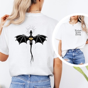 May include: White t-shirts with black graphic designs. One shirt features a dragon with outstretched wings and a sunburst design. The other shirt has the text "Violent Little Thing" in a gothic font.