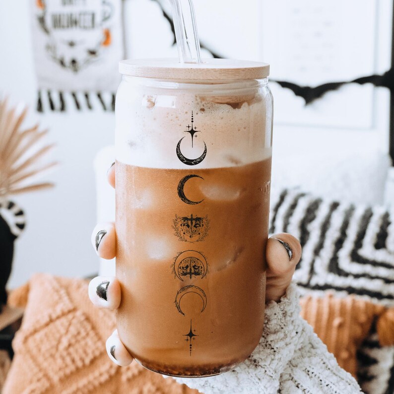May include: A clear glass tumbler with a wooden lid and a straw. The tumbler has a design of moons and stars on it. The tumbler is filled with a brown liquid.
