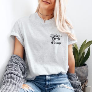 May include: A light gray t-shirt with the text "Violent Little Thing" in a gothic font. The shirt is made of a soft material and is suitable for casual wear. The shirt is paired with blue jeans and a gray sweater.