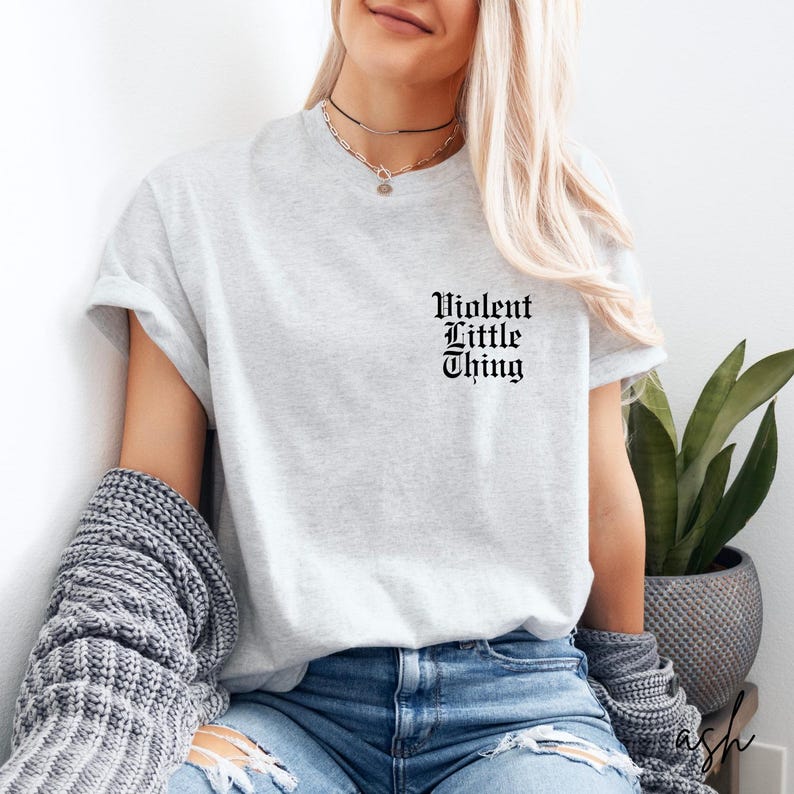 May include: A light gray t-shirt with the text "Violent Little Thing" in a gothic font. The shirt is made of a soft material and is suitable for casual wear. The shirt is paired with blue jeans and a gray sweater.