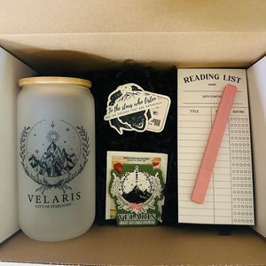 Pode incluir: Uma caixa de presente contendo um copo de vidro fosco com um design de montanha e o texto "VELARIS CITY OF STARLIGHT", adesivos, um bloco de notas de lista de leitura e um marcador rosa. A caixa é preenchida com papel preto.