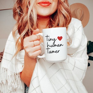 May include: White ceramic mug with black text that reads "Tiny human tamer" and a red heart. The mug is being held by a person wearing a white sweater.