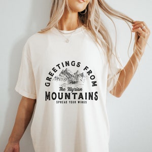 Puede incluir: Una camiseta blanca con un diseño gráfico negro que dice "Greetings from The Illyrian Mountains Spread Your Wings". El gráfico presenta una cordillera con una cabaña en primer plano.