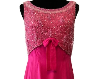 Puede incluir: Un vestido fucsia sin mangas, largo hasta el suelo. El corpiño está adornado con cuentas brillantes y un lazo a juego. El vestido se exhibe en un maniquí negro, mostrando su elegante diseño.