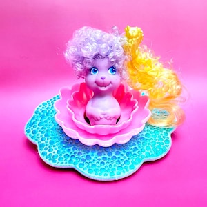Puede incluir: Una pequeña muñeca morada con cabello rizado color lavanda y una larga coleta amarilla, sentada dentro de una base de plástico rosa con forma de loto. La base descansa sobre una plataforma circular turquesa, festoneada y con textura.