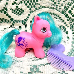 May include: Pink My Little Pony with a blue and turquoise mane and tail. The pony has a purple symbol on its side of a teddy bear wearing a hat. A purple comb is also included.