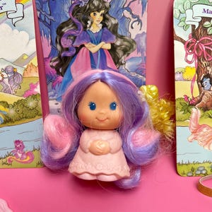 May include: A vintage doll with pink and purple hair, wearing a pink dress, is centered in front of illustrated book covers. The doll has blue eyes and a smiling expression. The book covers feature illustrations of a woman in a purple dress and a tree with pink ribbons.