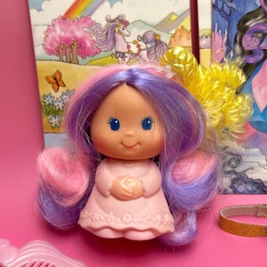 May include: A small doll with a peach-colored face, blue eyes, and long, wavy hair in shades of purple and pink. The doll wears a light pink dress with a floral pattern at the hem and has a bright yellow, curly hair accessory attached to the side.