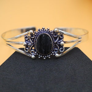 May include: A silver-toned cuff bracelet with an oval, black stone centerpiece. The stone is set in an ornate, filigree-style frame. The bracelet has a triple-band design on each side of the stone.