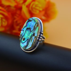 Puede incluir: Un anillo de plata con una incrustación ovalada de concha de abulón. La concha tiene un patrón de color verde, azul e iridiscente.