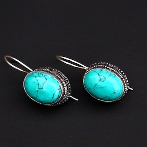 May include: A pair of silver earrings featuring oval turquoise stones. The stones are a vibrant blue-green colour with dark veining. The earrings have a decorative silver border and a simple hook closure. The background is black.