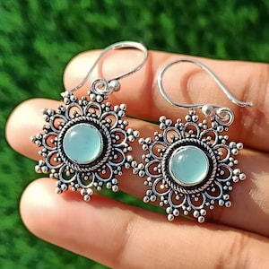Peut inclure: Une paire de boucles d'oreilles argentées avec une pierre précieuse ronde bleu clair au centre. Les boucles d'oreilles ont un motif floral avec un bord perlé et une fermeture à crochet. Elles sont tenues dans une main.