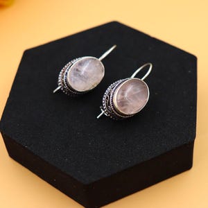 May include: A pair of silver earrings with oval-shaped pink rose quartz stones. The earrings have intricate silver designs around the stones.