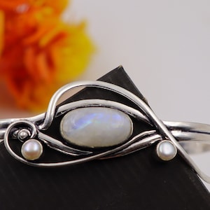 May include: A silver bracelet with a white moonstone and two pearl accents. The bracelet has a delicate, intricate design.
