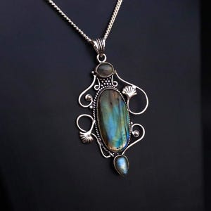 Könnte beinhalten: Silberfarbene Halskette mit Anhänger, verziert mit einem großen, ovalen Labradoritstein mit blau-grünen Schimmern. Der Anhänger hat ein verziertes Filigran-Design mit kleineren Steinen und einer zarten Kette. Die Halskette wird vor dunklem Hintergrund präsentiert.