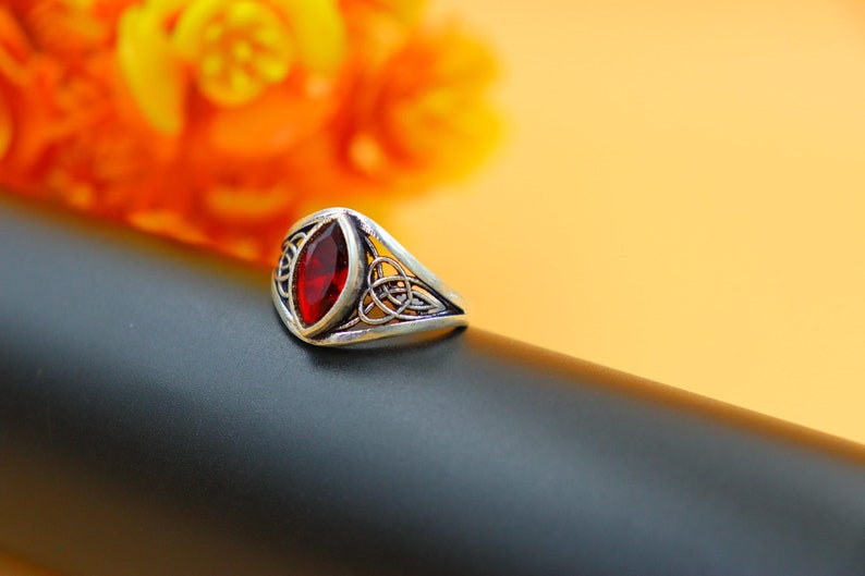 May include: A silver ring with a red gemstone set in a Celtic knot design. The ring is on a black surface.