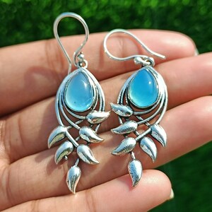 Peut inclure: Boucles d'oreilles pendantes en argent avec une pierre précieuse bleu clair au centre et un motif de feuilles délicates.