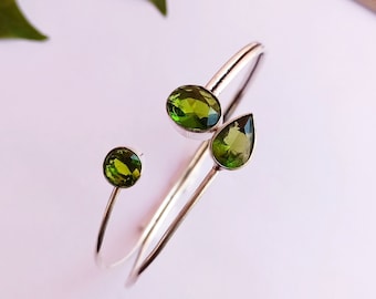 May include: Silver bangle bracelet featuring three green gemstones. The bracelet is open and has a simple design.