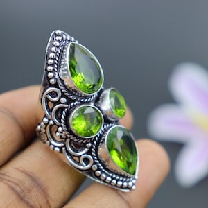 May include: A silver ring with an ornate design and four green gemstones. The ring is set on a finger.