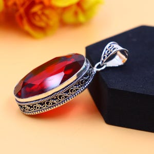 May include: A silver pendant with an ornate design features a large, oval-shaped red gemstone.