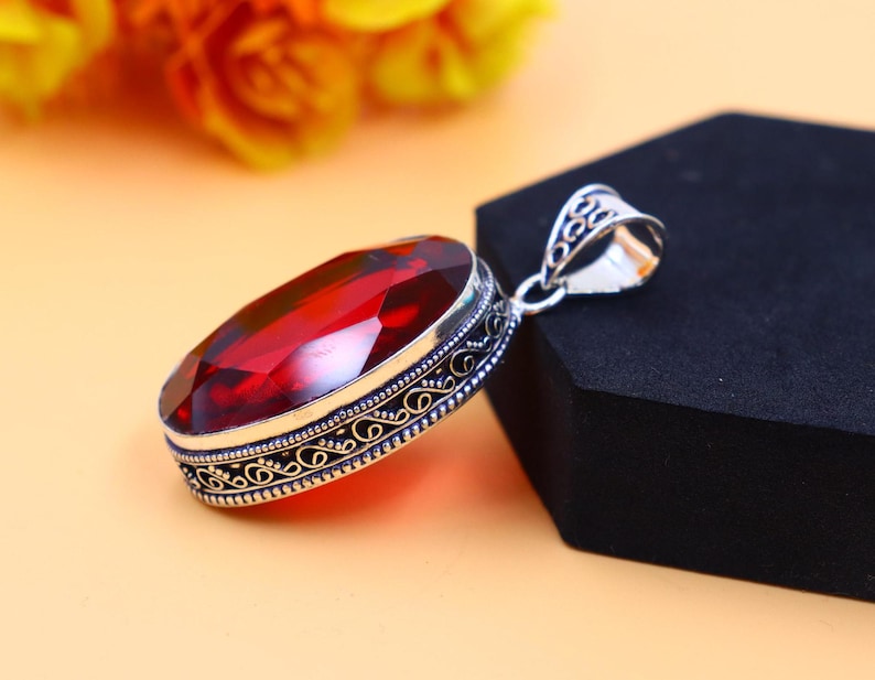 May include: A silver pendant with an ornate design features a large, oval-shaped red gemstone.