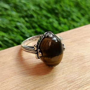 May include: A silver ring featuring a large oval tiger's eye gemstone. The ring has an intricate ornate design surrounding the stone.