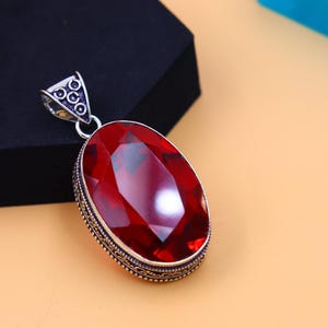 May include: A silver pendant featuring an oval-shaped red gemstone set within an intricate silver frame. The pendant has a silver bail for attaching to a chain.