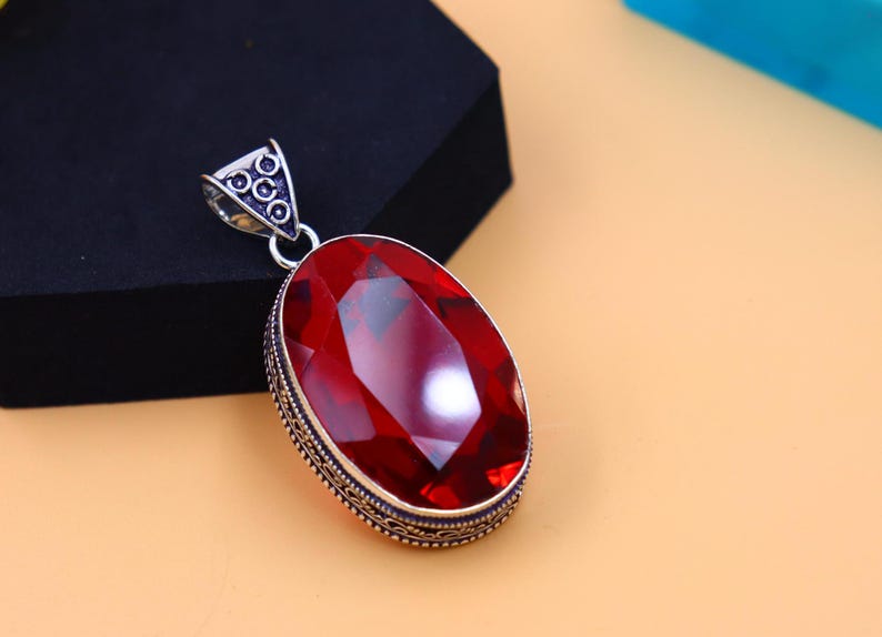May include: A silver pendant featuring an oval-shaped red gemstone set within an intricate silver frame. The pendant has a silver bail for attaching to a chain.