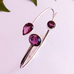 May include: A silver bangle bracelet featuring three purple gemstones. The bracelet is open and has a simple design.