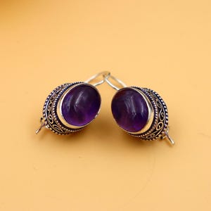 Puede incluir: Un par de pendientes de plata con piedras preciosas de amatista de forma ovalada. Los pendientes tienen intrincados detalles de plata alrededor de las piedras preciosas.