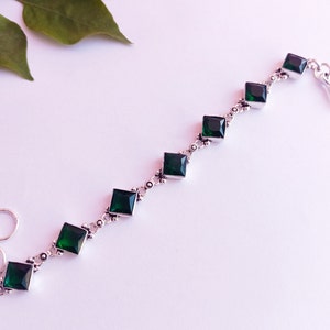 Puede incluir: Un brazalete de plata con una cadena y piedras preciosas cuadradas verdes.
