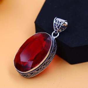 May include: A silver pendant with an ornate design features a large, oval-shaped red gemstone.