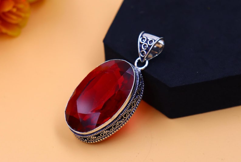 May include: A silver pendant with an ornate design features a large, oval-shaped red gemstone.