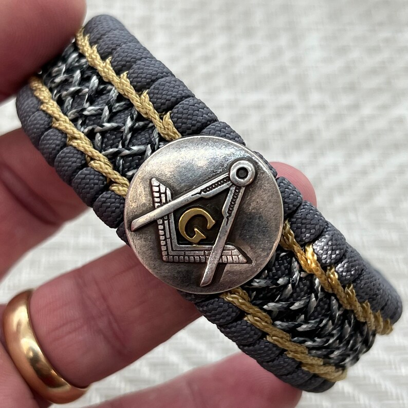 May include: A gray and gold paracord bracelet with a silver metal charm featuring a compass and the letter "G".