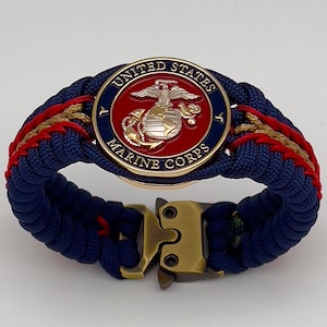 May include: A paracord bracelet featuring a circular United States Marine Corps emblem with a red center, gold eagle, globe, and anchor, surrounded by a blue border. The band is woven with navy blue, red, and tan cord and has a brass-toned metal buckle.