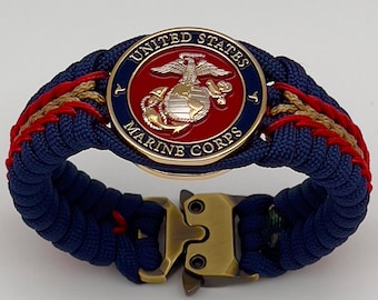 Puede incluir: Una pulsera de paracord con un emblema circular del United States Marine Corps con centro rojo, águila, globo terráqueo y ancla dorados, rodeados por un borde azul. La banda está tejida con cordón azul marino, rojo y beige y tiene una hebilla metálica.