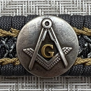 May include: A silver and gold Masonic compass and square with a letter G in the center. The compass and square are on a black and gold braided background.