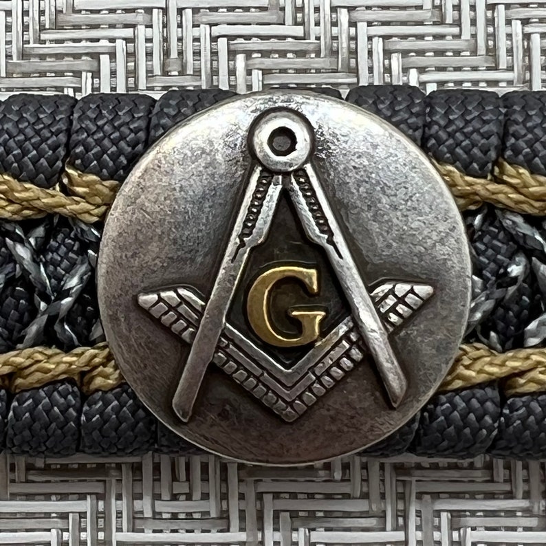 May include: A silver and gold Masonic compass and square with a letter G in the center. The compass and square are on a black and gold braided background.
