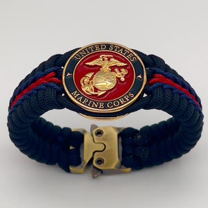 May include: A paracord bracelet featuring a central circular emblem with a gold eagle, globe, and anchor on a red background, surrounded by a black border with the text UNITED STATES MARINE CORPS. The band is woven with dark blue and red cord.