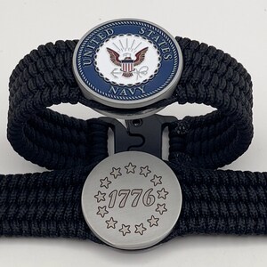 Op de afbeelding: Twee zwarte paracord armbanden. Eén armband heeft een cirkelvormig embleem met de tekst "UNITED STATES NAVY" en een adelaar. De andere armband heeft een cirkelvormig embleem met het nummer "1776" omringd door sterren.