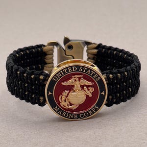 Może przedstawiać: Bransoletka z paracordu z czarno-beżowym plecionym paskiem i metalową klamrą. Okrągły emblemat pośrodku przedstawia logo United States Marine Corps z czerwonym tłem, złotym orłem, globusem i kotwicą oraz czarną obwódką.