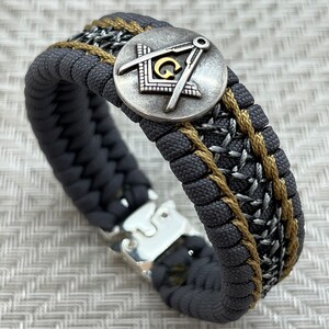 May include: A gray and gold braided bracelet with a silver metal charm featuring a compass and square design. The charm has a gold letter "G" in the center.