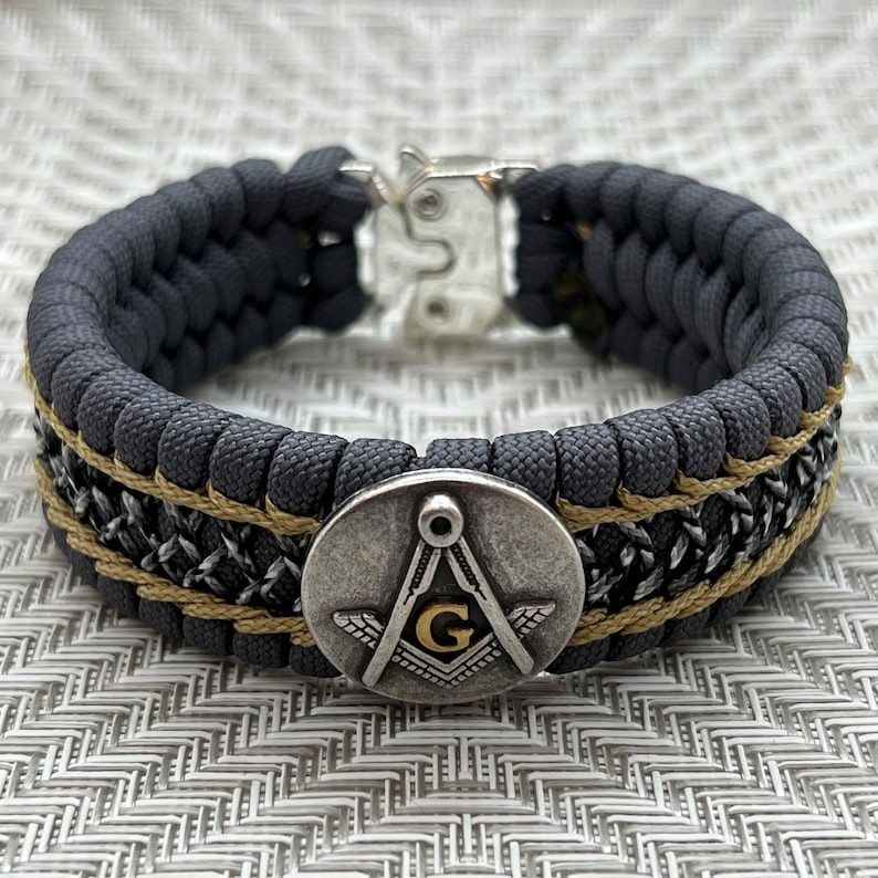 May include: A black paracord bracelet with a silver and gold Masonic symbol charm. The bracelet has a black and silver woven pattern and a tan accent.