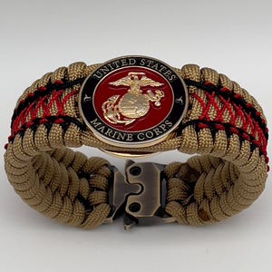 May include: A paracord bracelet featuring a tan woven cord with red and black accent stitching. A circular metal emblem is centered on the band, displaying the gold United States Marine Corps eagle, globe, and anchor logo on a red background.
