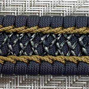 May include: Close-up of a black and gold braided trim with a diamond pattern. The trim is made of woven fabric and rope.