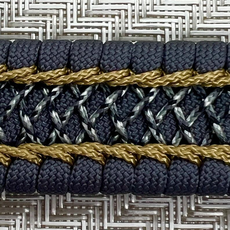May include: Close-up of a black and gold braided trim with a diamond pattern. The trim is made of woven fabric and rope.